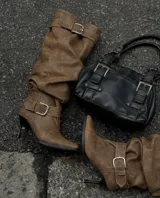 Botas de caña alta color camel, confeccionadas en material tipo gamuza, con diseño slouchy y puntera fina. Presentan dos correas con hebillas metálicas, una en la parte superior de la caña y otra en el tobillo, y cuentan con un taco fino.