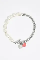 Collar de diseño asimétrico que combina una mitad de perlas irregulares y una mitad de cadena de eslabones metálicos plateados. Incluye un cierre de barra en T y dos dijes colgantes en forma de corazón, uno metálico con logo grabado y otro esmaltado en color rosado.