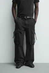 Pantalón cargo de corte relajado en color negro con efecto lavado. Presenta bolsillos laterales tipo cargo con solapas y cintas ajustables, bolsillos frontales y traseros, y detalles de argollas metálicas. Cierre frontal con cremallera y botón.