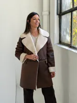 Abrigo de cuero sintético color marrón con interior y solapas de corderito sintético color beige. Presenta cierre frontal con botones, mangas largas con puños vueltos y bolsillos laterales.