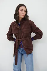 Chaqueta de gamuza en tono marrón oscuro, con cuello alto, cierre frontal con cremallera y botones a presión, bolsillos frontales con solapa y cinturón ajustable en la cintura.