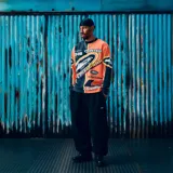 Buzo de manga larga Corteiz con diseño gráfico estilo motocross en colores naranja, negro y amarillo. Presenta múltiples estampados con logotipos y tipografías en el frente y las mangas.