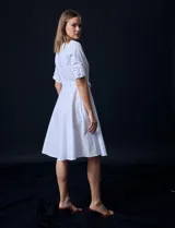 Vestido camisero blanco de algodón con cuello con solapa, delantera abotonada, mangas 3/4 regulables con presilla y botón, y cintura con lazo de ajuste.
