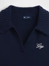 Buzo tipo polo de tejido de punto, color azul marino, con cuello solapa, escote en V y logo de la marca bordado en el pecho. Presenta mangas largas y un diseño de ruedo asimétrico con detalle acanalado.