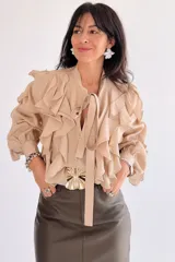 Camisa color camel con volados verticales en el frente y mangas largas abullonadas con puños ajustados. Presenta un lazo decorativo en el cuello y cierre frontal con botones.