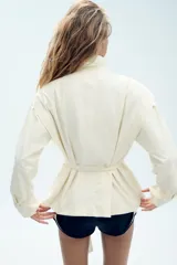Blazer corto de color marfil con cuello subido, manga larga y bolsillos laterales. Cuenta con un cinturón del mismo tejido para anudar en la cintura, forro interior a tono y cierre frontal con cremallera y botones a presión ocultos bajo solapa.