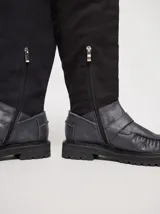 Bota híbrida de caña alta confeccionada en lona negra en la parte superior y cuero negro en la base tipo mocasín, con plataforma, suela track y tira con hebilla metálica cruzada en el empeine.