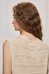 Chaleco de punto color beige, sin mangas, con escote redondo y cierre frontal con botones carey. Presenta diseño calado y silueta amplia.