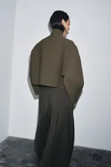 Chaqueta corta de tejido grueso color verde oliva, con cuello alto y cierre frontal completo de cremallera plateada. Presenta un corte amplio y mangas largas caídas.