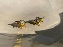 Par de aros dorados con forma de sol y cristal circular en el centro.