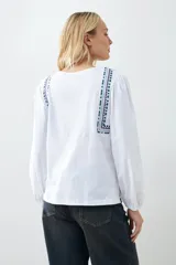 Remera blanca de manga larga con detalles bordados en tonos azules y verdes en el pecho y hombros. Presenta un escote en V con cordones ajustables terminados en borlas y puños elásticos.