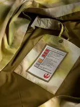 Poncho impermeable de diseño técnico con estampado camuflado en tonos tierra, cuello alto con cierre frontal parcial, capucha integrada y cinturón ajustable con compartimentos. Incluye parches decorativos en el pecho.