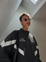 Campera deportiva negra con cuello alto, cierre frontal completo y franjas blancas en las mangas. Presenta estampados en color rosado en el pecho, incluyendo la inscripción "Only F.A.N.S." y gráficos de palmeras con estrellas.