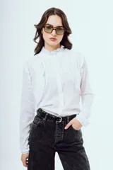 Camisa blanca de manga larga con cuello alto con volados, detalles de alforzas y pliegues en el frente y cierre frontal con botones ocultos.