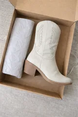 Bota estilo texana de caña media, color blanco hueso, con taco cuadrado de madera y costuras decorativas en forma de alas en la caña.