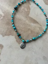 Gargantilla de cuentas de cristal en tonos tornasolados, violetas y oscuros, con dije central ovalado de metal plateado con diseño de escudo.