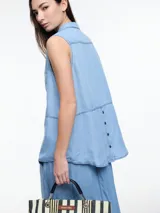 Camisa sin mangas en denim 100% lyocell, muy liviano con caída fluida. De corte recto y largo medio, presenta cartera escondida en el frente y un bolsillo plaqué en el pecho. En la espalda, un detalle distintivo: botones decorativos alineados en abertura vertical. Los pespuntes a contratono completan el look.