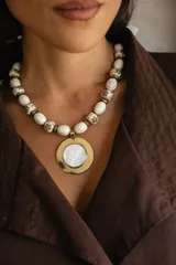 Collar corto con cuentas de piedra reconstituida veteada en tonos crema, intercaladas con cuentas metálicas tibetanas grabadas. Presenta un dije central circular con borde metálico dorado y centro de nácar.