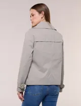 Gabardina corta gris, de silueta ligeramente entallada con diseño clásico de trench, cuello camisero con solapas amplias, cierre cruzado con doble botonadura y bolsillos laterales diagonales.