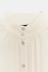 Blusa fluida de color blanco roto o crema, con cuello alto fruncido y manga larga con puños elásticos tipo nido de abeja. Presenta un frente abotonado con botones pequeños y brillantes.