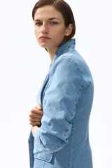 Blazer de denim de algodón celeste, con solapas de muesca, hombreras y mangas tres cuartos fruncidas. Cuenta con bolsillos delanteros con solapa y diseño abierto sin cierre.