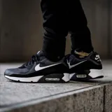 Championes Nike Air Max 90 365, diseño clásico con unidad de aire visible en el talón, confeccionados en una combinación de malla y materiales sintéticos en color negro con detalles en blanco.