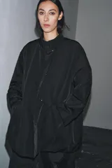 Chaqueta oversize de nylon en color negro, con cuello redondo subido, manga larga y cierre frontal con botones a presión ocultos. Presenta bolsillos delanteros y forro interior a tono.