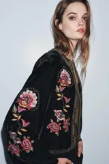 Chaqueta abierta de terciopelo negro con cuello redondo y mangas largas acampanadas. Presenta bordados florales en tonos rosados y verdes, con aplicaciones de abalorios y detalles dorados en el cuello y la parte frontal.