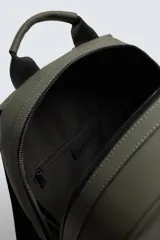 Mochila de color verde militar con textura grabada, diseño minimalista y forma redondeada. Cuenta con un compartimento principal y un bolsillo frontal, ambos con cierre de cremallera. Posee asa de mano y asas de hombro acolchadas y ajustables.
