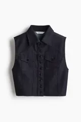 Chaleco corto de denim de algodón rígido, color azul oscuro, con cuello, cierre frontal de botones de metal y dos bolsillos superiores con solapa y botón. Presenta un corte ajustado con canesú y costuras visibles que aportan estructura.