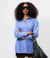 Camisa alargada de manga larga, confeccionada en tricolina de algodón con diseño de rayas finas azules y blancas. Cuenta con cuello inglés, cierre frontal con botones y un bolsillo aplicado en el pecho.