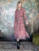 Vestido camisero midi de gasa estampada con flores rojas y blancas sobre fondo marrón. Tiene cuello con solapa, abertura central con botones, mangas largas y lazo de ajuste en la cintura.