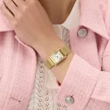 Reloj analógico de cuarzo con caja rectangular de acero inoxidable en tono dorado. Presenta una esfera blanca con números romanos y está adornado con cristales en los laterales de la caja. La correa es de acero inoxidable tipo malla milanesa en tono dorado.