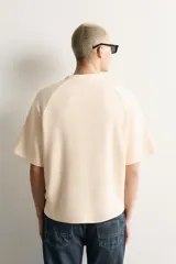 Camiseta relaxed fit de color beige, con cuello redondo, mangas cortas y estampado frontal en tono azul con texto gráfico.
