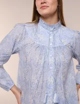 Blusa celeste de algodón con estampado floral, cuello mao fruncido con volado y cordel frontal con borlas. Tiene mangas largas abullonadas con puños elásticos y abotonadura frontal.