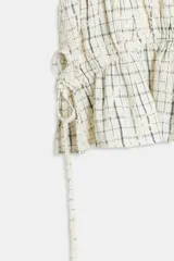 Blusa de manga corta abullonada con diseño a cuadros, cuello con volante y acabados elásticos. Cuenta con aberturas y lazos ajustables en los laterales.