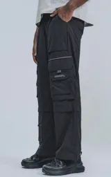Pantalón cargo negro de silueta oversize, confeccionado en tejido técnico liviano de poliéster. Presenta múltiples bolsillos cargo con solapa y detalles reflectivos horizontales en la parte superior de los bolsillos.