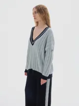 Buzo tejido de punto con diseño oversized, cuello en V profundo y mangas largas. Presenta un patrón de trenzas verticales en color verde oscuro con detalles en contraste de color rosado en el cuello, puños y ruedo.