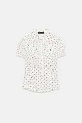 Top de popelina de color blanco con estampado de lunares negros, cuello mao con escote en pico y lazada, mangas cortas abullonadas y cierre frontal con botones.