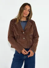 Chaqueta de pana color marrón con diseño corto, cuello con solapas deshilachadas, cierre frontal con botones y dos bolsillos delanteros con solapa.