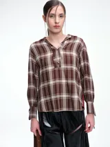 Camisa de corte relajado con estampado de cuadros en tonos marrones y beige. Presenta cuello camisero, cierre frontal parcial con botones y mangas largas con puños ajustables mediante lazos. El diseño destaca por un panel de encaje en la espalda que aporta contraste.