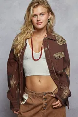 Chaqueta de algodón color marrón con diseño estilo patchwork, que incorpora parches con estampado animal print de leopardo en hombros y bolsillo frontal. Presenta detalles de tachas metálicas, bordes deshilachados, cierre frontal con botones metálicos y un acabado con efecto de pintura desgastada en las mangas.