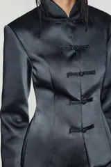 Chaqueta negra de tejido satinado, con cuello mao, manga larga con hombreras, corte evasé y cierre frontal con alamares.