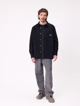 Camisa de gabardina color verde oliva, con cuello clásico, cierre frontal de botones y dos bolsillos frontales con parche. Incluye un pequeño parche con el logo de la marca en el bolsillo izquierdo.