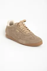 Zapatilla deportiva de gamuza color visón, con cordones a tono y suela de goma marrón. Presenta un diseño retro de running con el nombre de la marca grabado en dorado en el lateral.