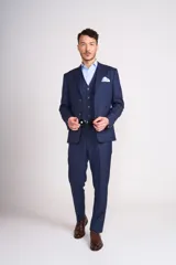 Traje de tres piezas color azul marino con textura sutil. Incluye saco entallado con solapas clásicas, chaleco abotonado y pantalón de corte recto. Se complementa con una camisa celeste de cuello italiano y pañuelo de bolsillo blanco.