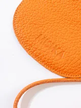 Monedero pequeño semicular de cuero vacuno texturizado color naranja, con cierre superior, tirador de correa de cuero y argolla lateral. Cuenta con el logotipo de la marca grabado en el frente.
