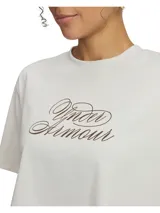 Remera corta (crop top) de manga corta, color blanco hueso, con el logo de Under Armour estampado en el pecho en tipografía cursiva de color marrón.