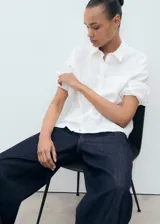 Camisa de lino blanca de corte recto con cuello camisero, manga larga con puños abotonados, cierre frontal de botones y un bolsillo de parche en el pecho.