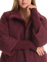 Tapado de tejido bouclé color marrón, con silueta evasé y largo a la cadera. Presenta cuello alto envolvente, cierre frontal oculto con botones a presión y bolsillos frontales de gran tamaño. Sus mangas son 3/4 con corte amplio y puños de tejido rib al tono que se extienden para cubrir las muñecas.